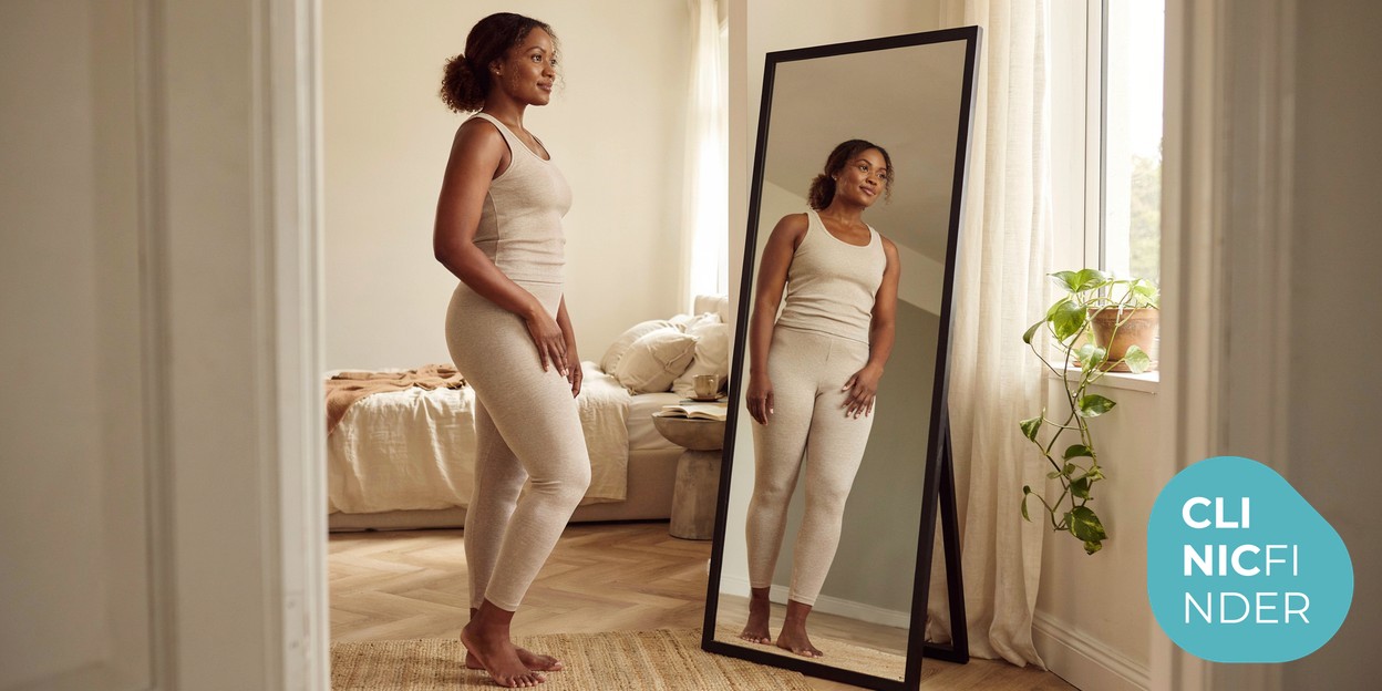 A woman standing side-on assessing her waist and flank silhouette in a full-length mirror in a bright room