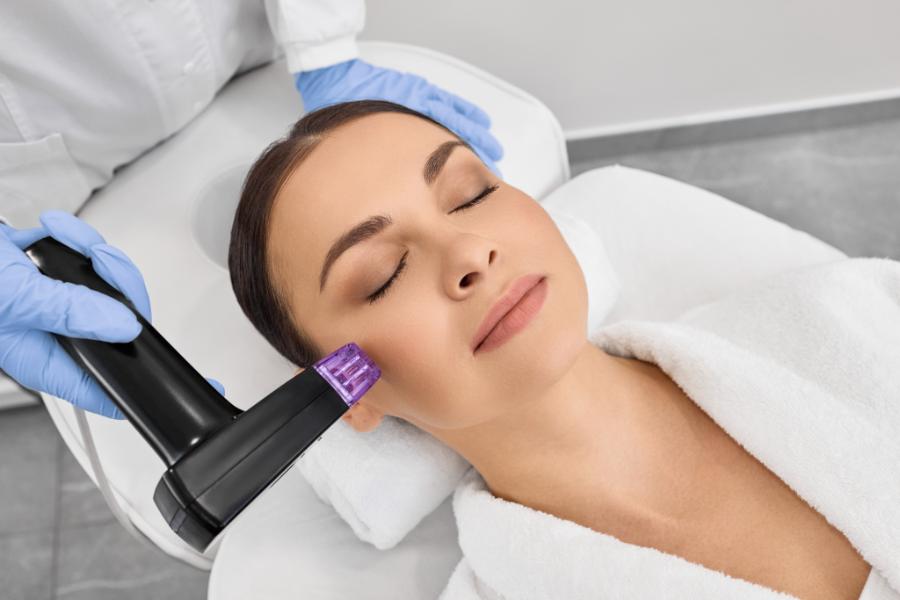Practitioner in blue gloves holding a radiofrequency microneedling handpiece with a square cartridge tip against a client’s cheek